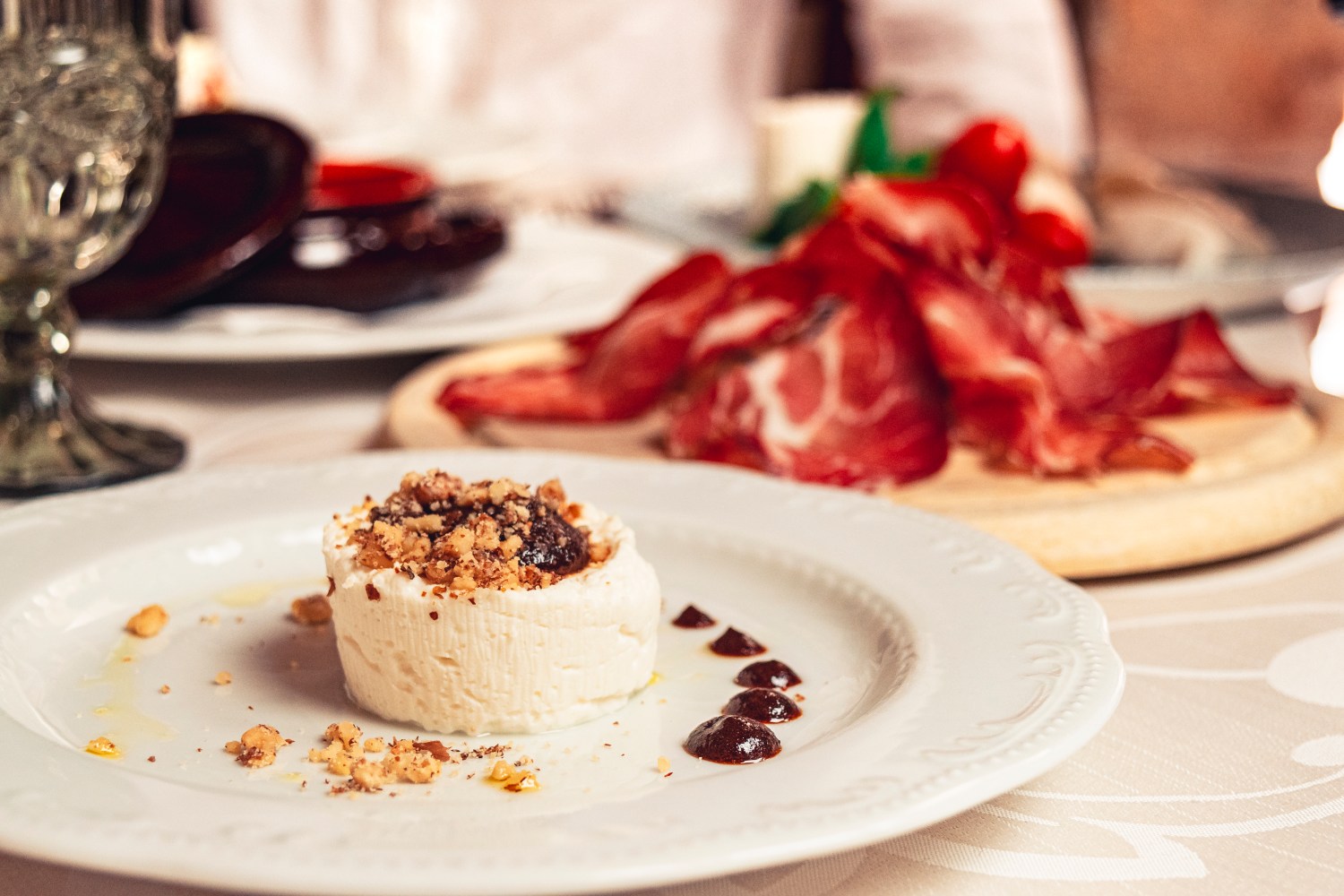 Cheese with nuts on plate, sliced cured meat on wooden board in the background.