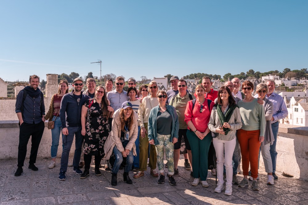 A group of 21 people posing outdoors on a sunny day in front of white buildings and a clear sky.