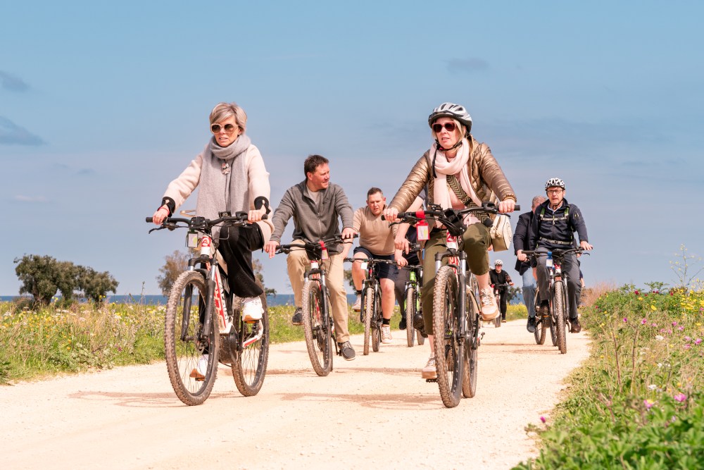 Group of people cycling on a sunny dirt path with flowers and a clear sky.