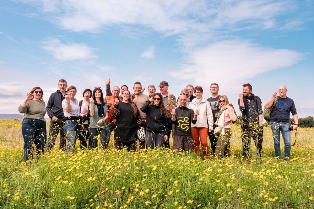 Group of 20 people smiling in a field of yellow flowers under a blue sky.