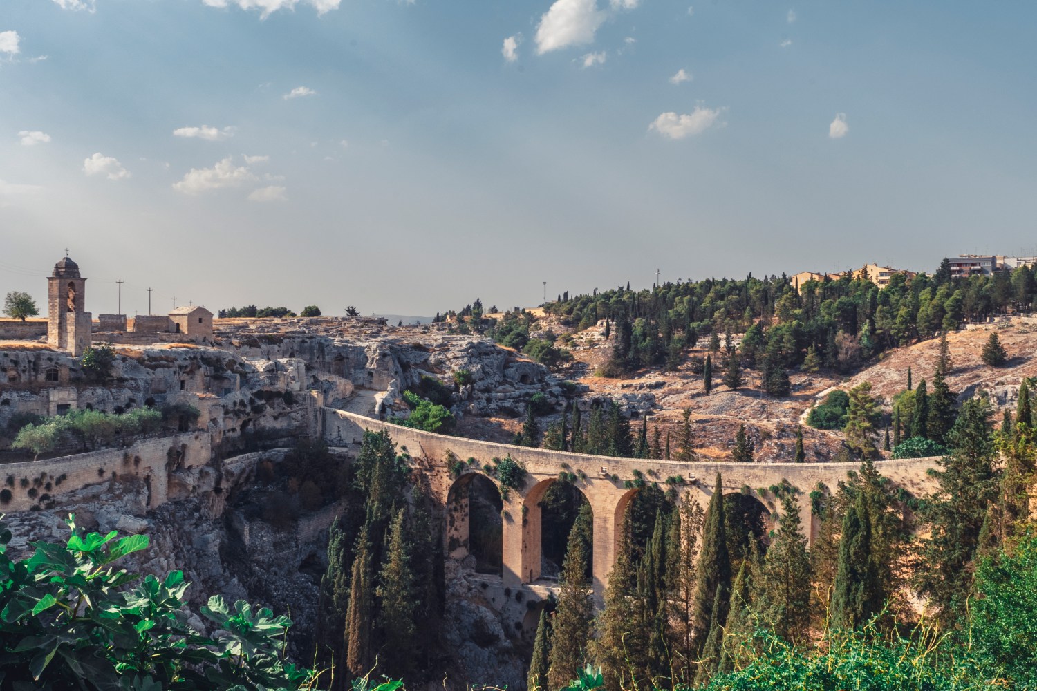 Stone bridge spans a forested valley with buildings on a rocky cliff under a blue sky.