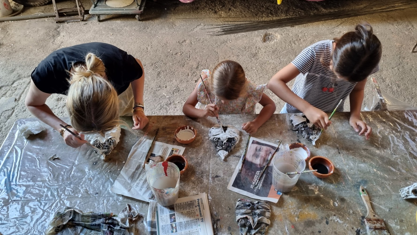 Three people painting crafts at a table covered with newspaper and art supplies.