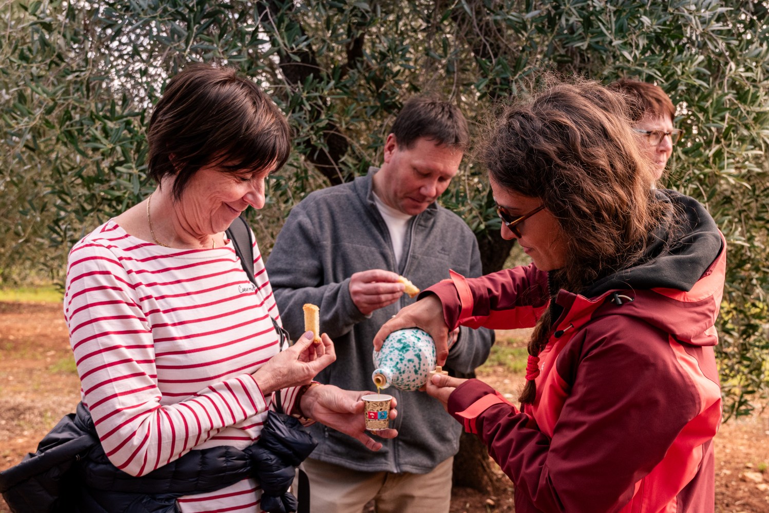 People tasting olive oil outdoors, one pours from a ceramic bottle, others hold bread pieces.
