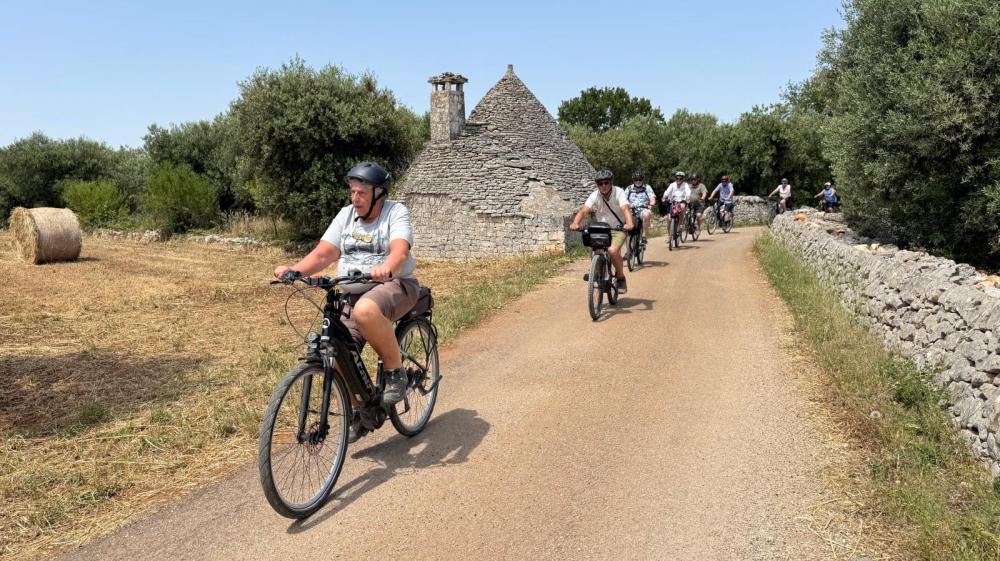 Cyclists ride on a rural path beside a trullo, with trees and hay bales surrounding.