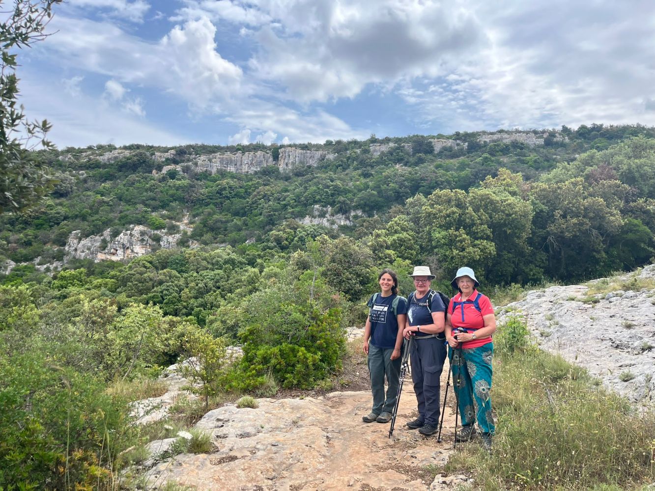 Three hikers with walking poles standing on a rocky trail, surrounded by lush green hills under a cloudy sky.