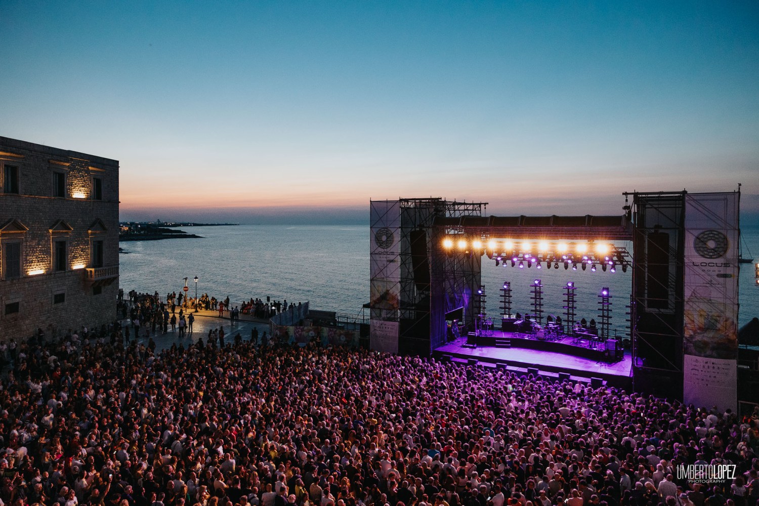 Outdoor concert by the sea at sunset with a large crowd and illuminated stage.